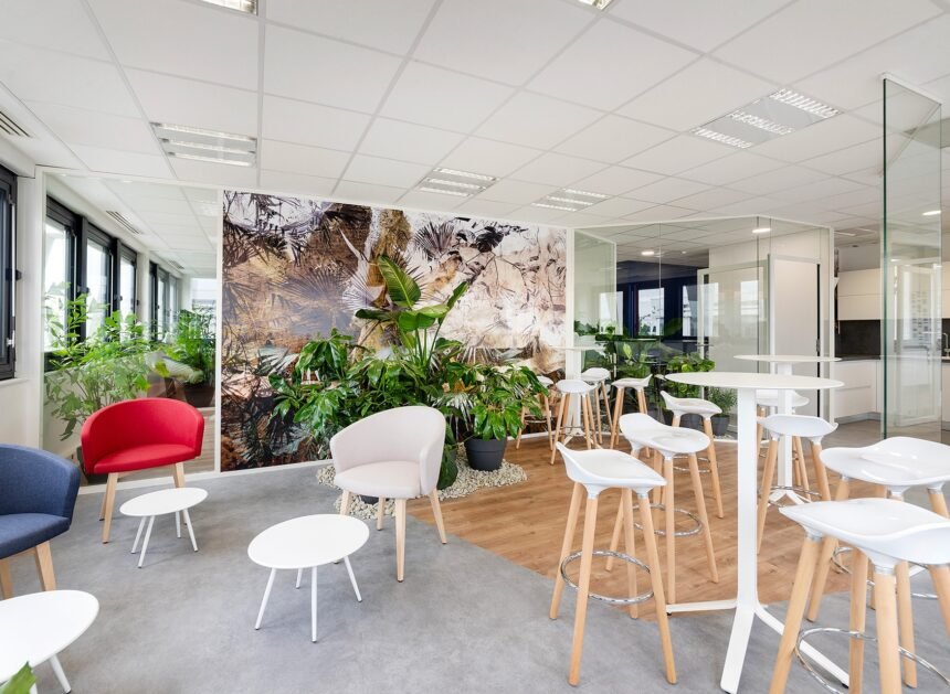Salon de bureau moderne avec des chaises et des tables blanches, des fauteuils colorés, des plantes d'intérieur et de grandes fenêtres laissant entrer la lumière naturelle.
