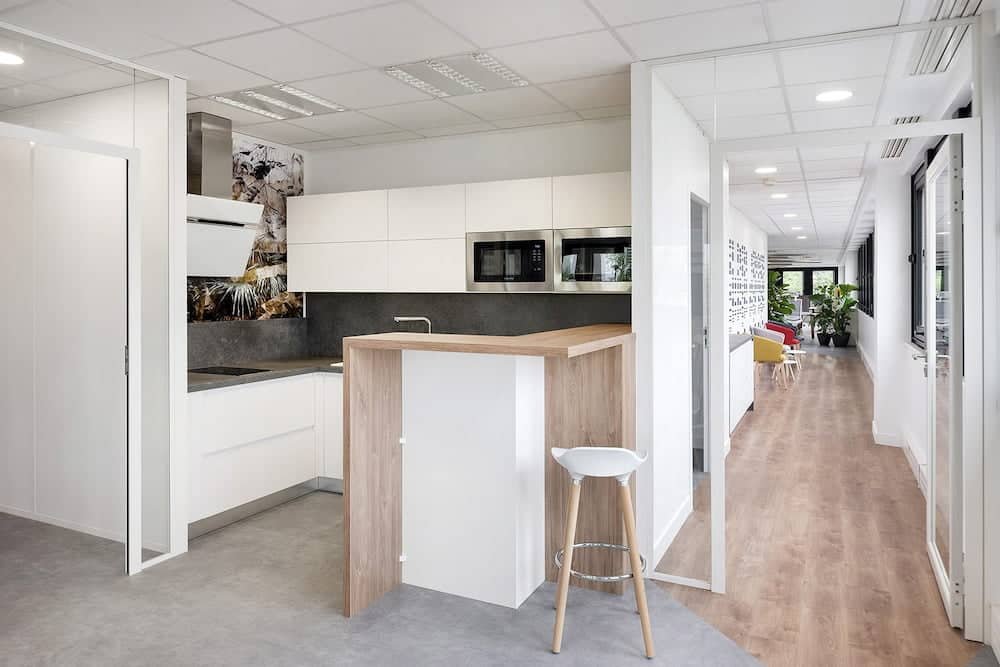 Kitchenette de bureau moderne avec des armoires blanches, des appareils encastrés, un petit bar de petit-déjeuner en bois et un tabouret blanc. Couloir avec sol en bois et coin salon visible à l'arrière-plan.
