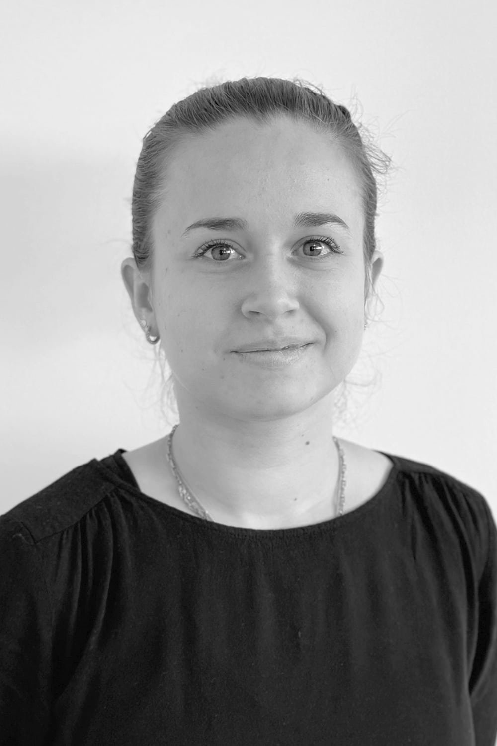 Une femme à la peau claire et aux cheveux attachés, portant un haut et un collier noirs, fait face à l'appareil photo sur un fond blanc uni.