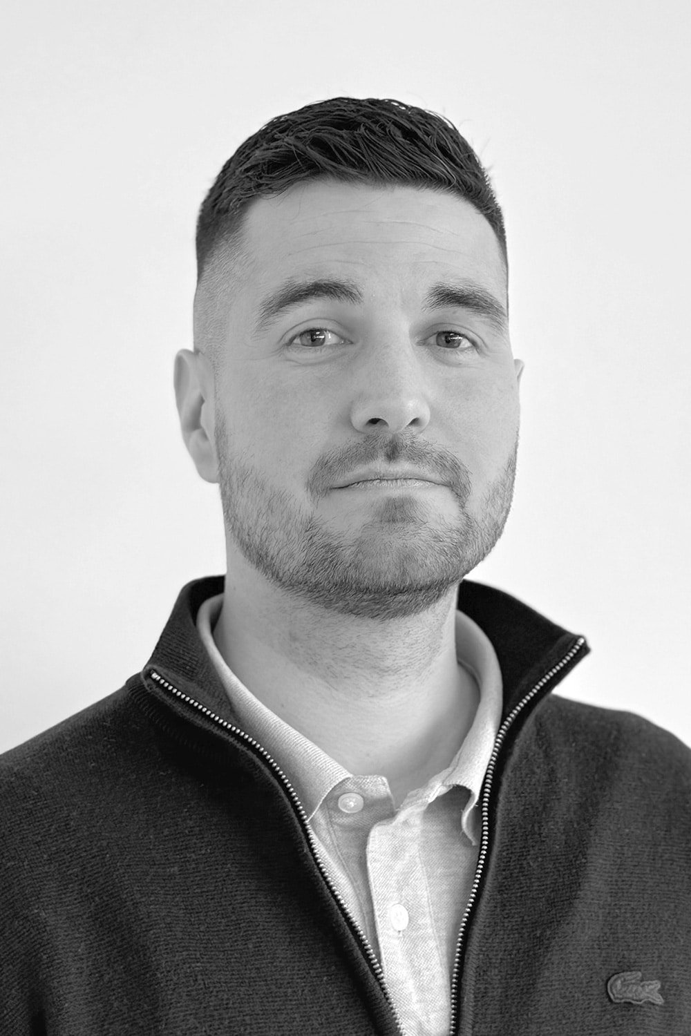 Un homme aux cheveux bruns courts et à la barbe taillée, portant une veste zippée sur une chemise à col, regarde l'appareil photo avec une expression neutre sur un fond uni.