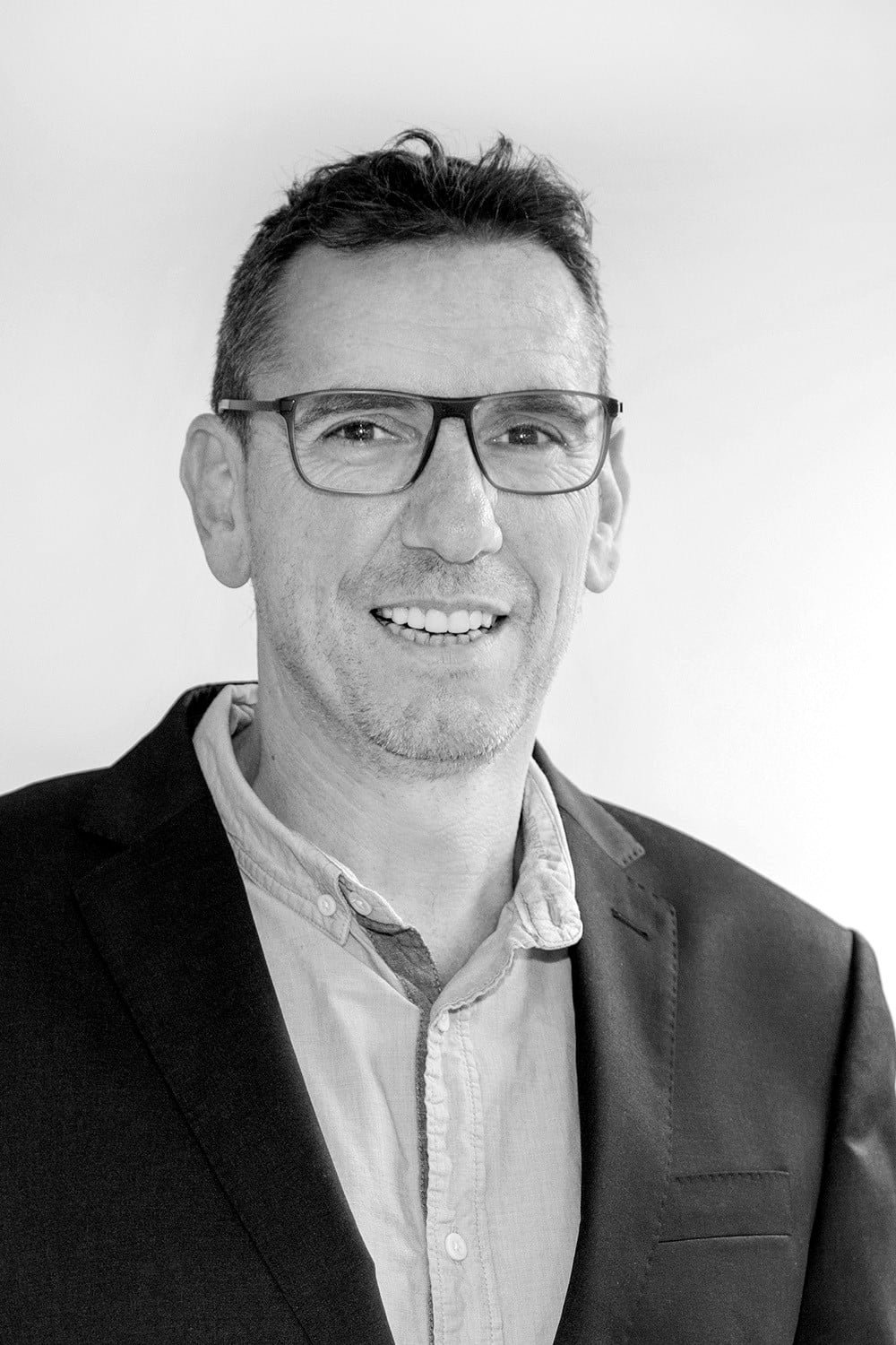 Un homme portant des lunettes, un blazer et une chemise à col, souriant et faisant face à l'appareil photo sur un fond uni.