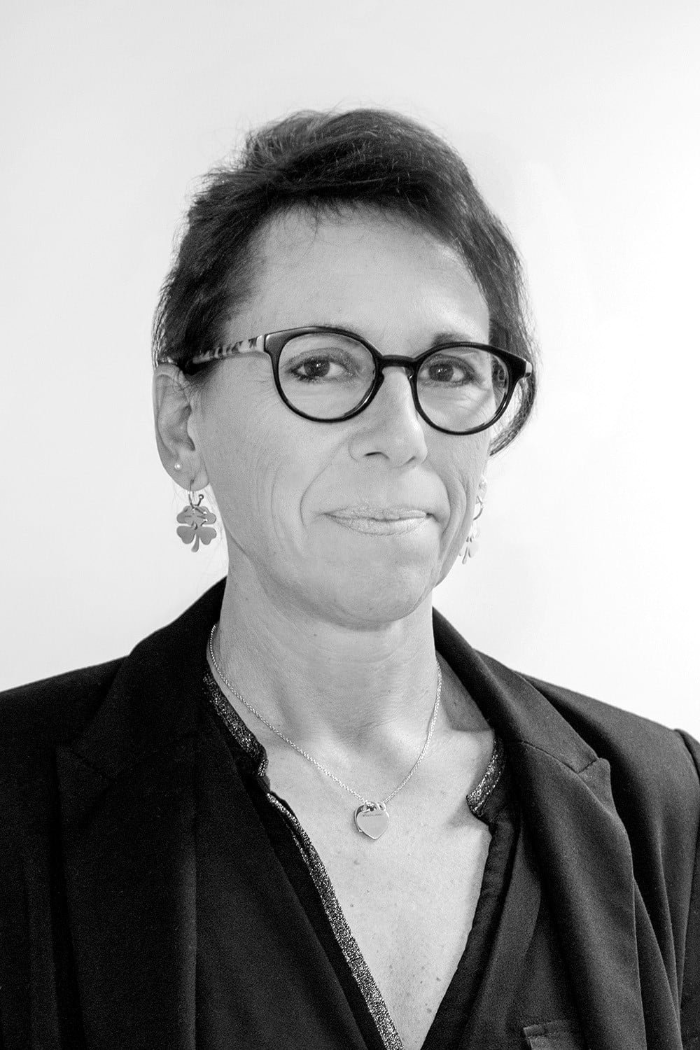 Une personne aux cheveux bruns courts, portant des lunettes et des boucles d'oreilles, vêtue d'un blazer et d'un collier, pose sur un fond uni.