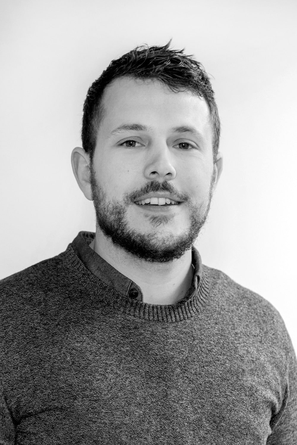 Un homme aux cheveux bruns courts et à la barbe, portant un pull sur une chemise à col, faisant face à l'appareil photo et souriant légèrement sur un fond uni.