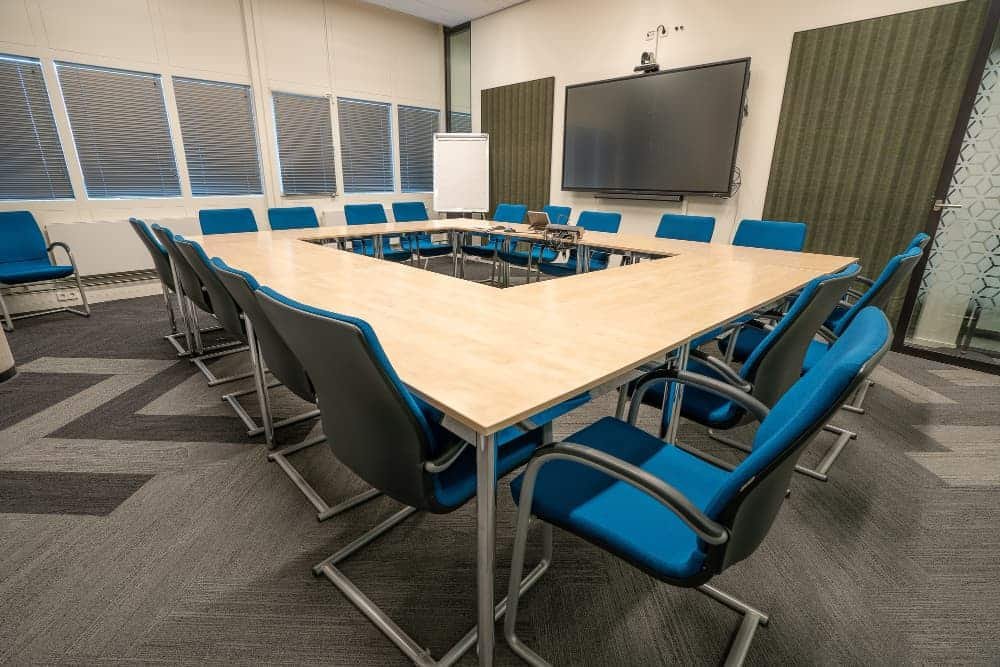 Une salle de conférence moderne avec une table en U, des chaises bleues, un grand écran mural, un tableau de conférence et des stores.