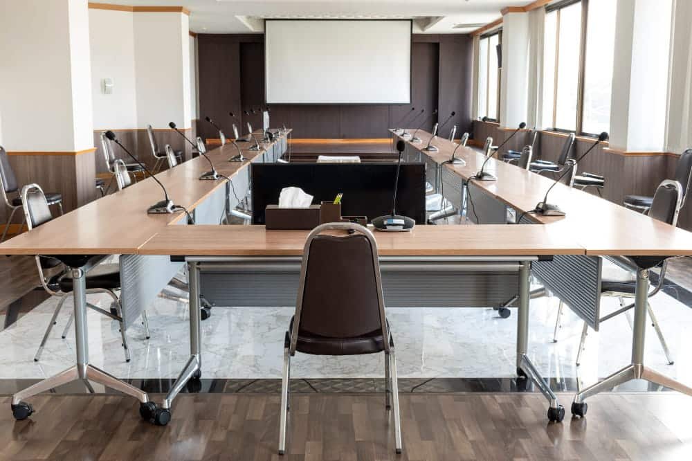 Salle de conférence vide avec une table en U, des microphones, des chaises, une boîte à mouchoirs et un grand écran de projection à l'avant.