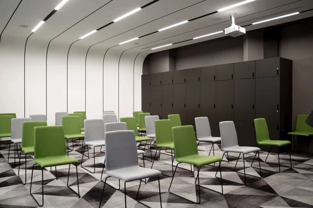 Une salle de conférence moderne avec des chaises vertes et grises disposées en rangées, une moquette géométrique, des casiers le long du mur du fond et un projecteur fixé au plafond.