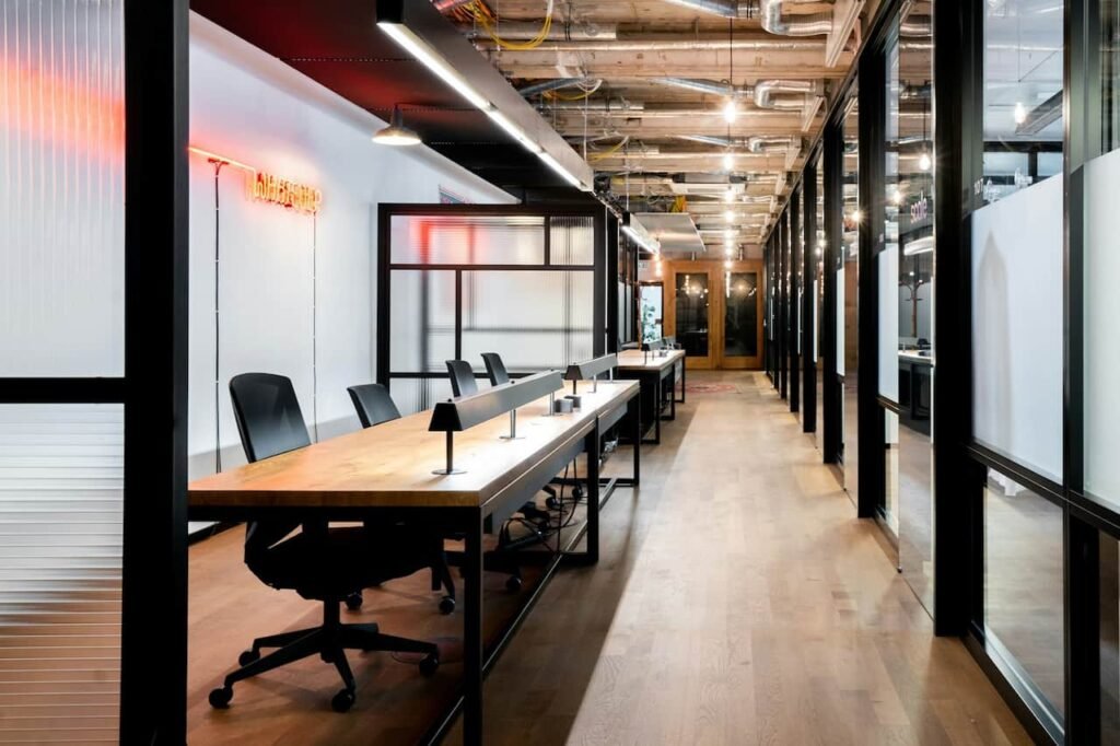 Bureau moderne à aire ouverte avec de longs bureaux partagés, des chaises noires, des lampes de bureau, des murs en verre et des tuyaux de plafond apparents, éclairés par un éclairage naturel et artificiel.