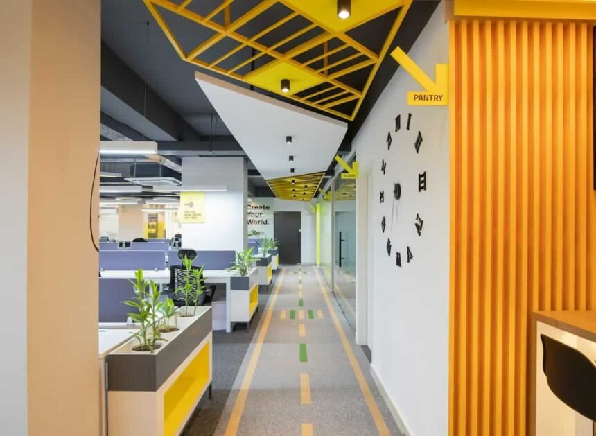 Couloir de bureau moderne avec un plafond géométrique, des plantes d'intérieur, un panneau directionnel de garde-manger et des cabines visibles sur la gauche.