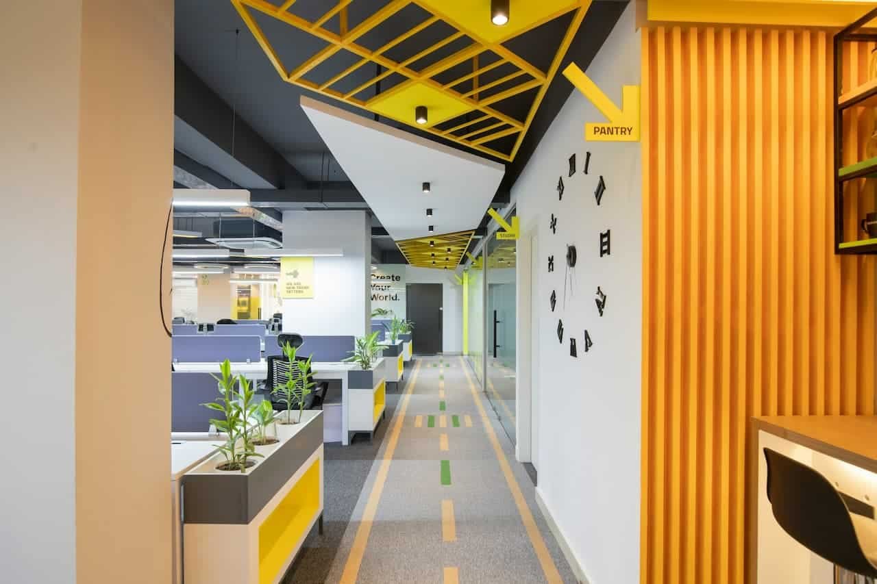 Couloir de bureau moderne avec un plafond géométrique, des plantes d'intérieur, un panneau directionnel de garde-manger et des cabines visibles sur la gauche.