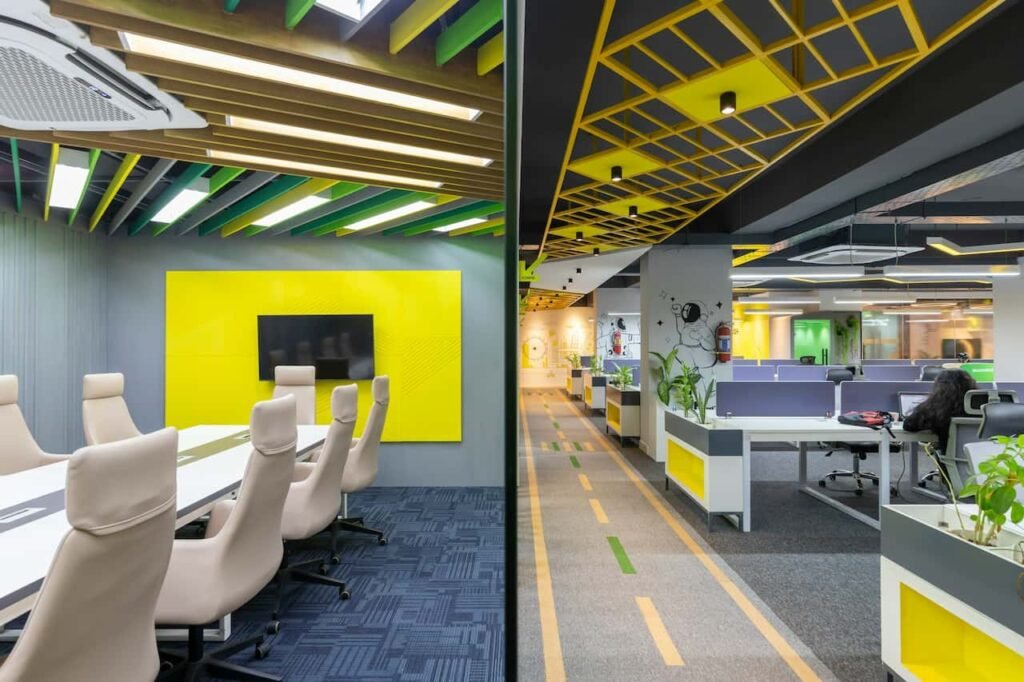 Intérieur de bureau moderne avec une salle de conférence dotée de chaises beiges et d'une télévision sur un mur jaune, et un espace de travail ouvert avec des cabines et des motifs géométriques au plafond.