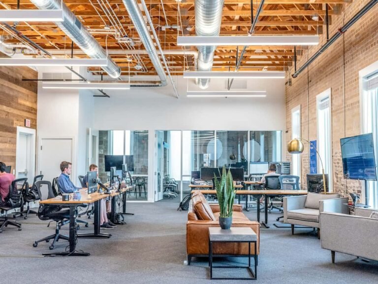 Bureau moderne à aire ouverte avec des personnes travaillant à des bureaux, un plafond apparent, une salle de réunion en verre, un coin salon avec canapé et de grandes fenêtres laissant entrer la lumière naturelle.