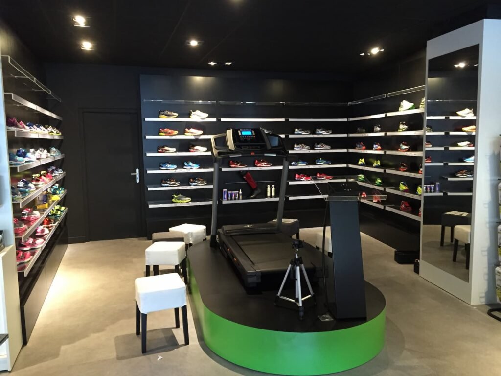 Un magasin de chaussures moderne présente des étalages muraux de chaussures de sport, un tapis roulant sur une plate-forme surélevée, des tabourets et un trépied dans un intérieur minimaliste et bien éclairé.