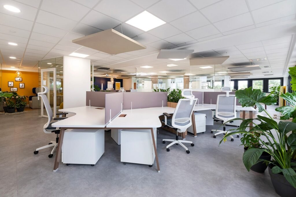 Bureau moderne à aire ouverte avec des bureaux blancs, des chaises ergonomiques, des panneaux de séparation, des plantes et de grandes fenêtres qui laissent passer la lumière naturelle. L'espace de travail est propre et organisé.