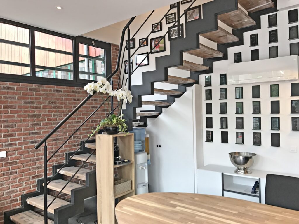 Intérieur moderne avec un escalier en bois, une rampe en métal noir, un mur en briques apparentes, des photos encadrées au mur et une petite étagère contenant une plante et une fontaine d'eau.