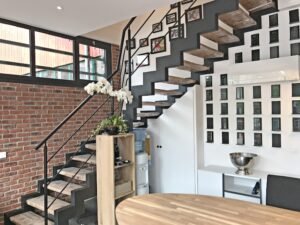 Intérieur moderne avec un escalier en bois, une rampe en métal noir, un mur en briques apparentes, des photos encadrées au mur et une petite étagère contenant une plante et une fontaine d'eau.
