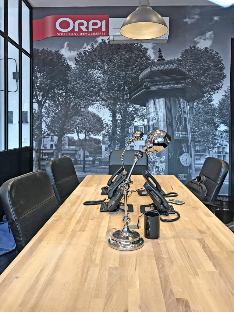 Une salle de réunion moderne avec une table en bois, des chaises noires, des téléphones de bureau, une lampe et une fresque de paysage urbain en noir et blanc avec le logo ORPI sur le mur.