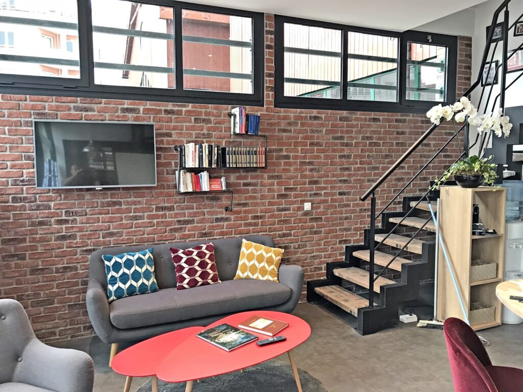 Salon moderne avec mur en briques apparentes, canapé gris avec coussins à motifs, télévision murale, petite bibliothèque, table basse rouge et escalier en métal noir à côté d'une plante en pot.