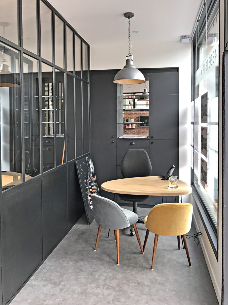 Petit bureau moderne avec une cloison en verre, une table ronde, trois chaises dépareillées, des armoires, une lampe suspendue et un téléphone sur la table.