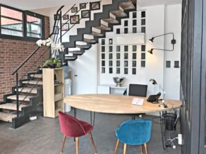 Bureau moderne avec un bureau en bois, des chaises colorées, des étagères murales et un escalier avec des photos encadrées au mur. Les briques apparentes et les blocs de verre sont visibles.