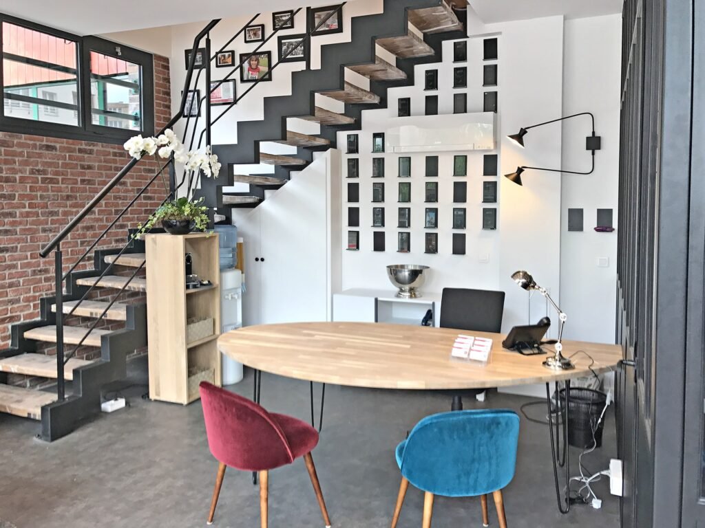 Un intérieur de bureau moderne avec un bureau en bois, des chaises colorées, un escalier en métal et des images encadrées au mur.