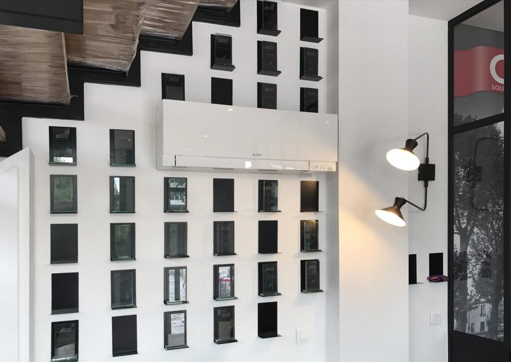 Un mur avec une grille de petites étagères rectangulaires noires, un climatiseur au centre et une double applique sur le côté droit.