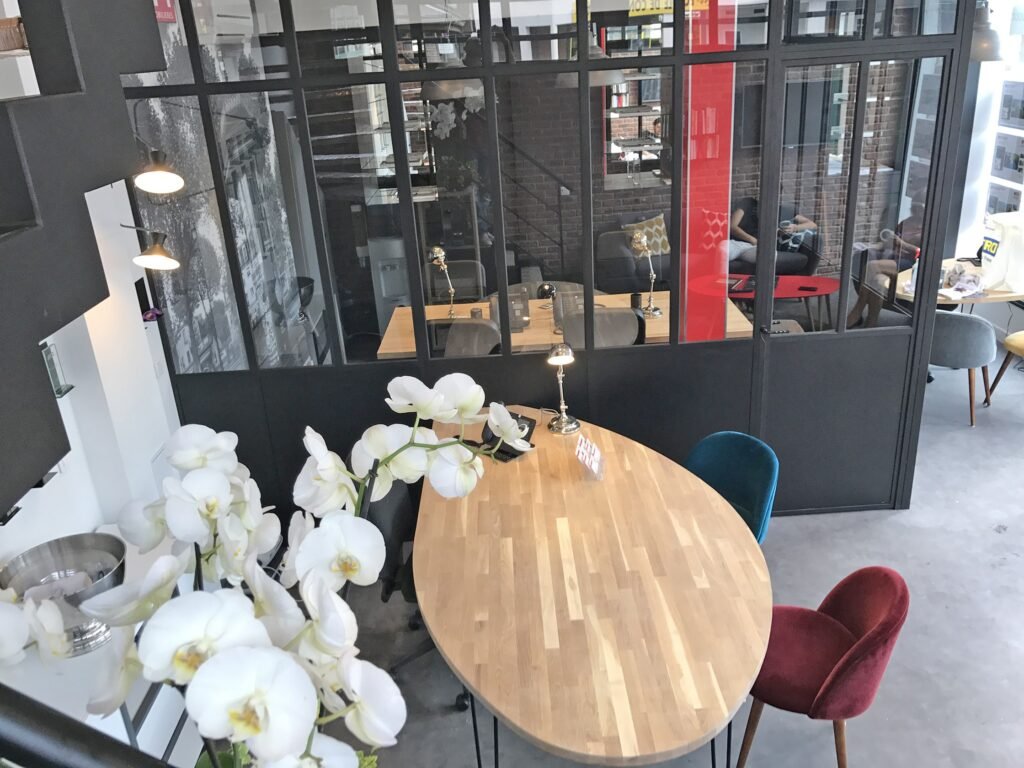 Bureau moderne avec une table ovale en bois, des chaises en velours coloré, des orchidées blanches au premier plan et des cloisons en verre séparant les zones de travail.