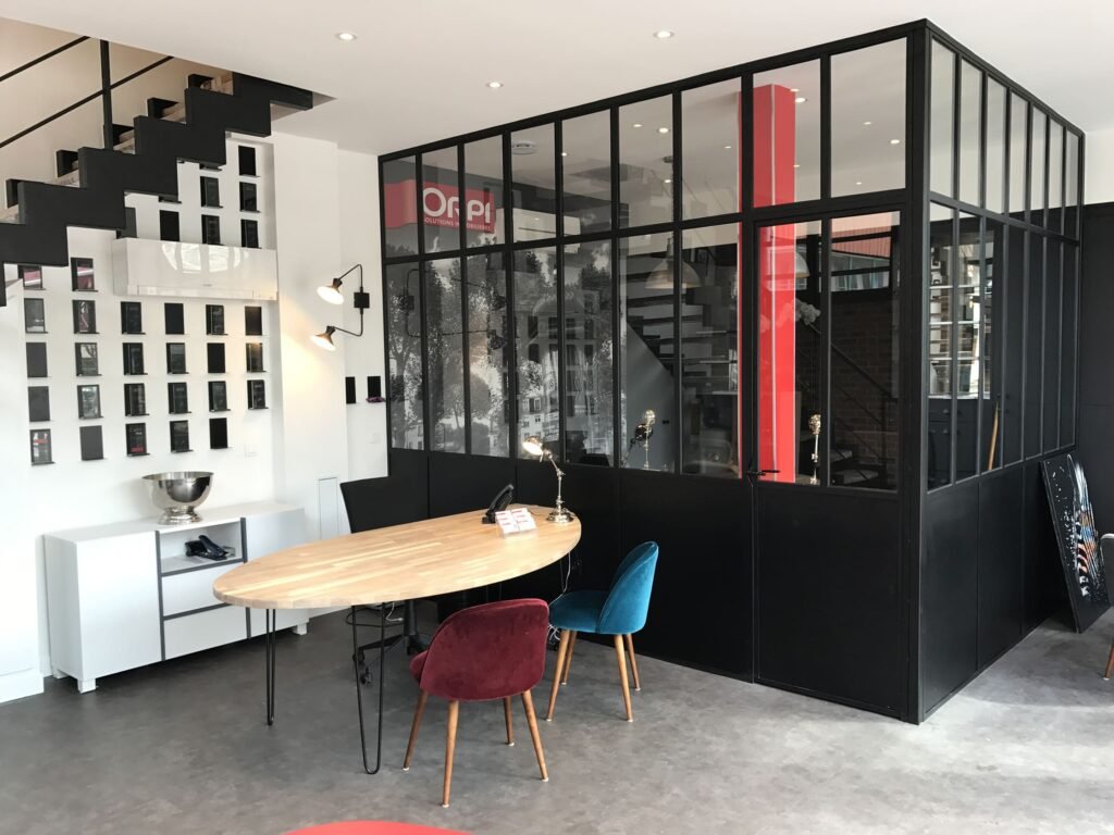 Bureau moderne avec un bureau en bois, des chaises colorées, une cloison en verre, un escalier et une décoration murale dans un espace ouvert et lumineux.