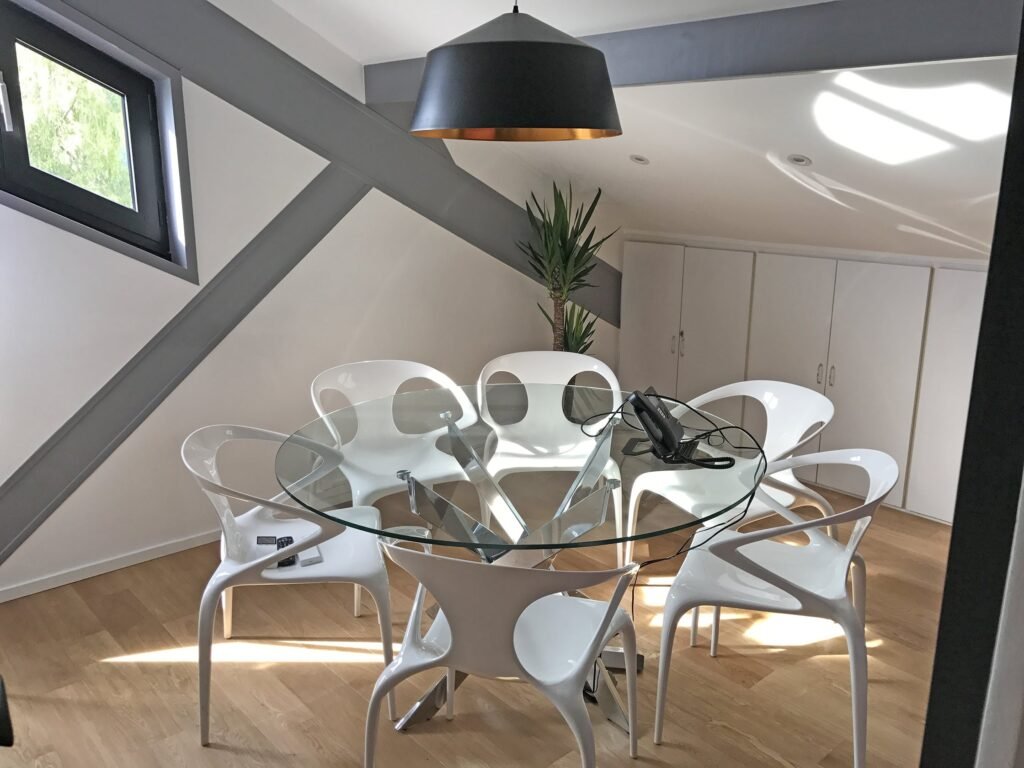 Salle de réunion moderne avec une table ronde en verre, six chaises blanches, une lampe suspendue noire, une plante en pot et des poutres inclinées au plafond.