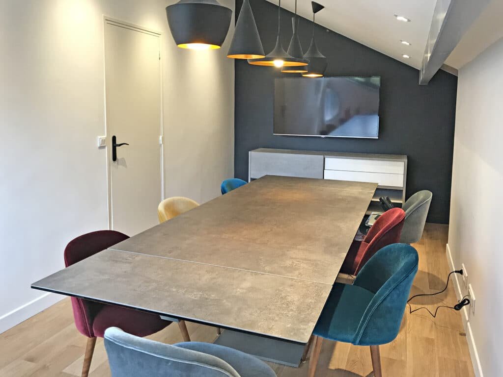 Salle de conférence moderne avec une longue table, des chaises en velours coloré, des lampes suspendues, une télévision murale et un buffet sur un sol en bois.