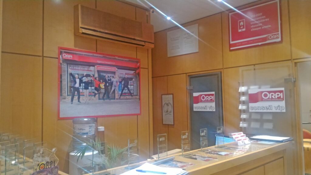 Réception d'un bureau avec des murs en bois, la marque ORPI, une grande photo sur le mur, des brochures sur le comptoir et des cloisons en verre portant la mention "conseil vip".