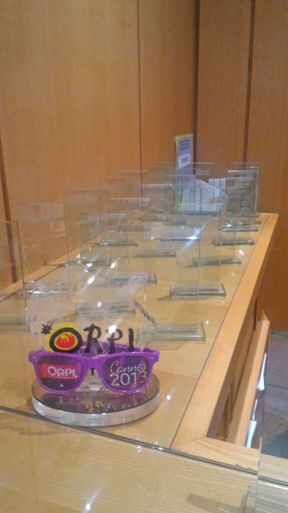 Une vitrine contenant plusieurs récompenses en verre transparent, avec un article décoratif coloré comportant des verres violets et le texte "2013" sur le devant.