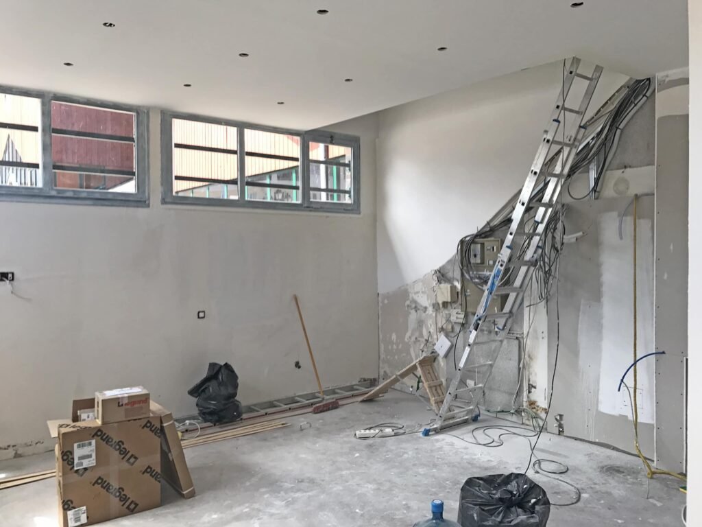 Pièce intérieure en cours de rénovation avec des câbles exposés, des échelles, des boîtes, des outils, des débris de construction, des murs et des plafonds non finis.