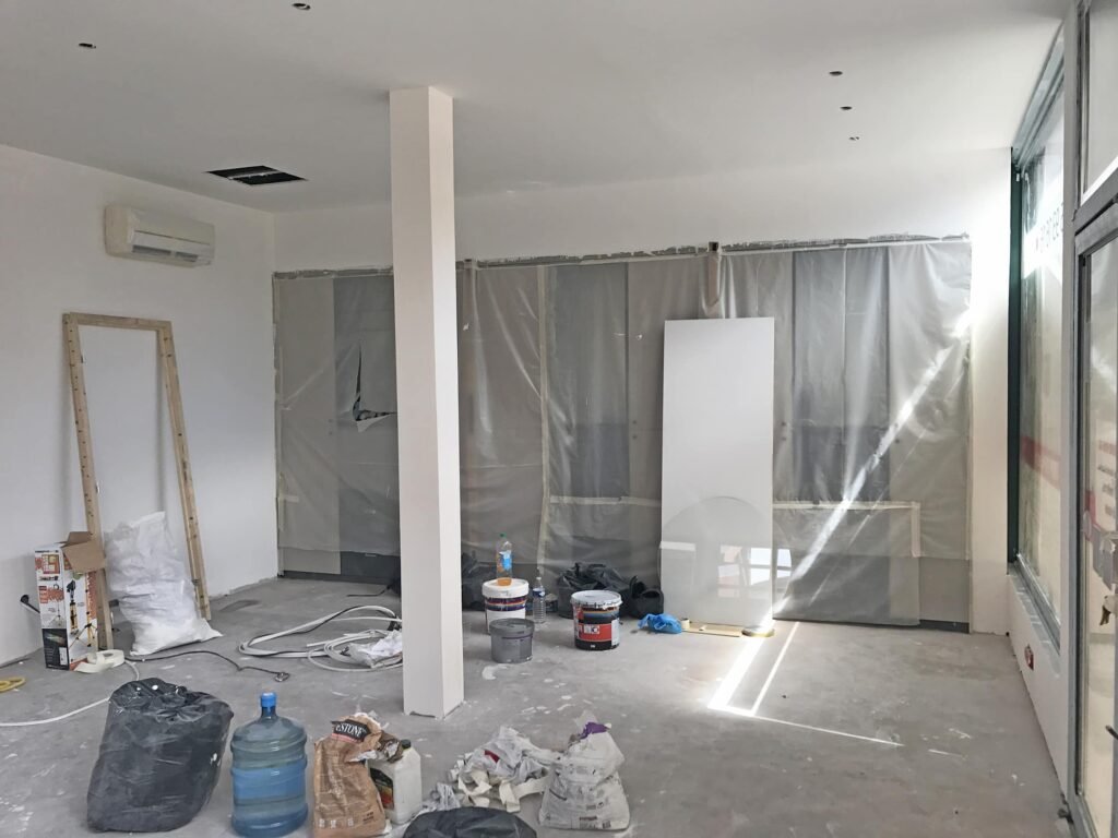 Une pièce en cours de rénovation avec des fenêtres recouvertes de plastique, des pots de peinture, des matériaux de construction et des outils éparpillés sur le sol.