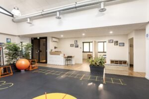 Clinique moderne de rééducation ou de thérapie physique avec des équipements d'exercice, des plantes, un bureau d'accueil et un coin salon sous un éclairage lumineux.