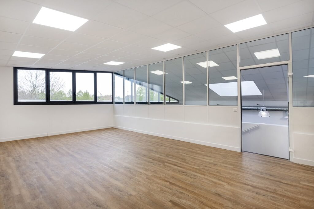 Espace de bureau vide et moderne avec sol en bois, grandes fenêtres, cloisons en verre et éclairage fluorescent.