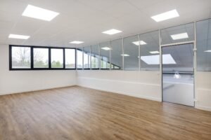 Espace de bureau vide et moderne avec sol en bois, grandes fenêtres, cloisons en verre et éclairage fluorescent.