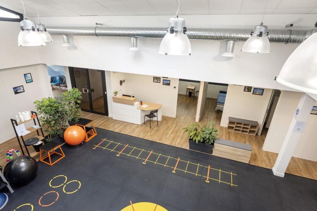 Salle de sport ou studio de remise en forme avec ballons d'exercice, échelles d'agilité marquées au sol, plantes en pot et bureau d'accueil. L'espace est doté d'un plancher en bois et d'un éclairage zénithal.