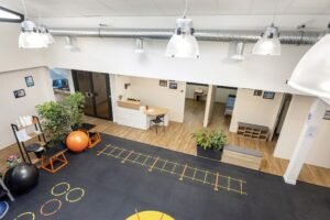 Salle de sport ou studio de remise en forme avec ballons d'exercice, échelles d'agilité marquées au sol, plantes en pot et bureau d'accueil. L'espace est doté d'un plancher en bois et d'un éclairage zénithal.