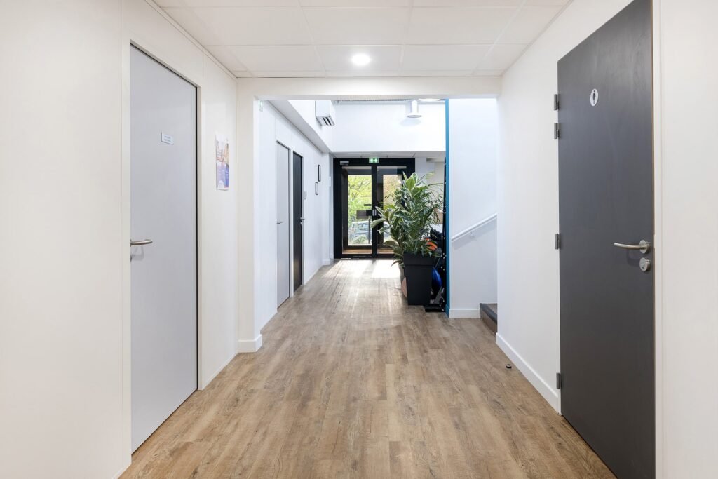Un couloir propre et moderne avec des murs clairs et un sol en bois, plusieurs portes fermées de part et d'autre, et une porte vitrée au bout menant à l'extérieur.