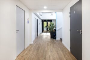 Un couloir propre et moderne avec des murs clairs et un sol en bois, plusieurs portes fermées de part et d'autre, et une porte vitrée au bout menant à l'extérieur.