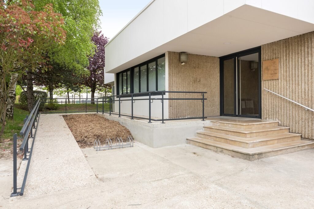 Une entrée de bâtiment moderne avec une rampe et des escaliers, des garde-corps en métal, de grandes fenêtres et un porte-vélos à l'avant. Des arbres et des aménagements paysagers entourent le patio.