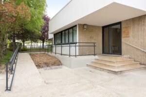 Une entrée de bâtiment moderne avec une rampe et des escaliers, des garde-corps en métal, de grandes fenêtres et un porte-vélos à l'avant. Des arbres et des aménagements paysagers entourent le patio.