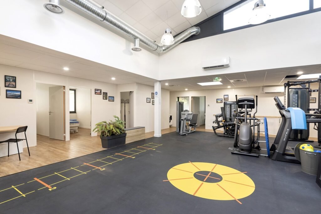 Salle de sport moderne dotée d'équipements d'exercice, d'une zone d'entraînement marquée au sol, de grandes fenêtres et d'un éclairage zénithal lumineux. Une plante en pot et des photos accrochées au mur décorent l'espace.