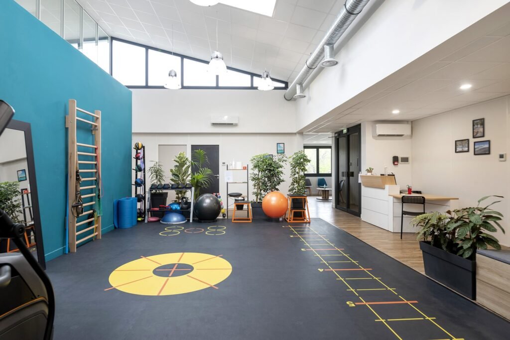 Salle de sport moderne équipée de matériel d'exercice, de ballons d'équilibre, d'une échelle murale, de marquages au sol pour les exercices d'agilité, de miroirs, de plantes et d'un bureau d'accueil dans un espace ouvert et lumineux.