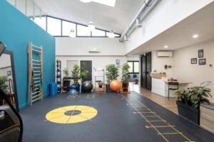 Salle de sport moderne équipée de matériel d'exercice, de ballons d'équilibre, d'une échelle murale, de marquages au sol pour les exercices d'agilité, de miroirs, de plantes et d'un bureau d'accueil dans un espace ouvert et lumineux.