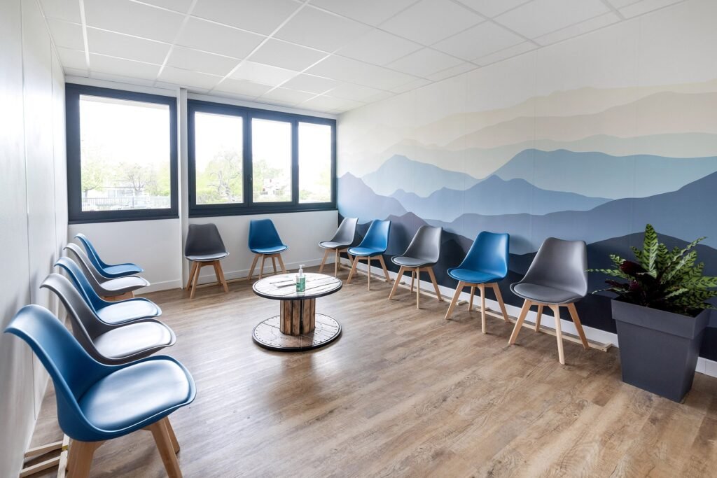 Une salle d'attente moderne avec des chaises bleues et grises disposées le long des murs, une table ronde au centre et une grande plante dans le coin. Un mur est orné d'une peinture murale représentant des montagnes abstraites.