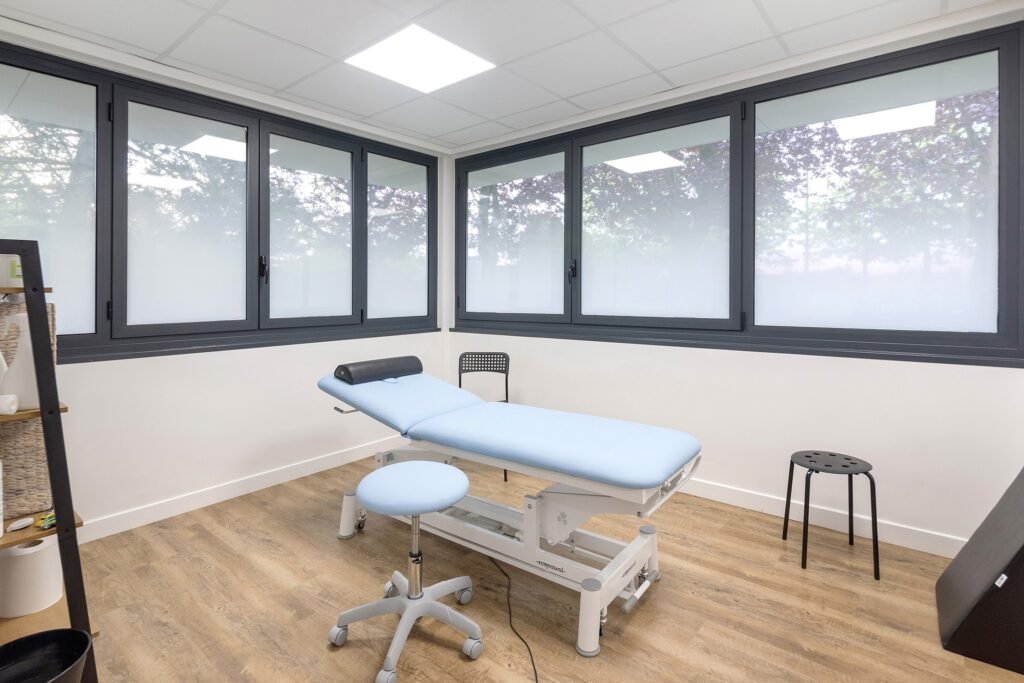 Une salle d'examen claire et propre avec une table d'examen bleu clair, un tabouret à roulettes et un petit tabouret noir, sous de grandes fenêtres.