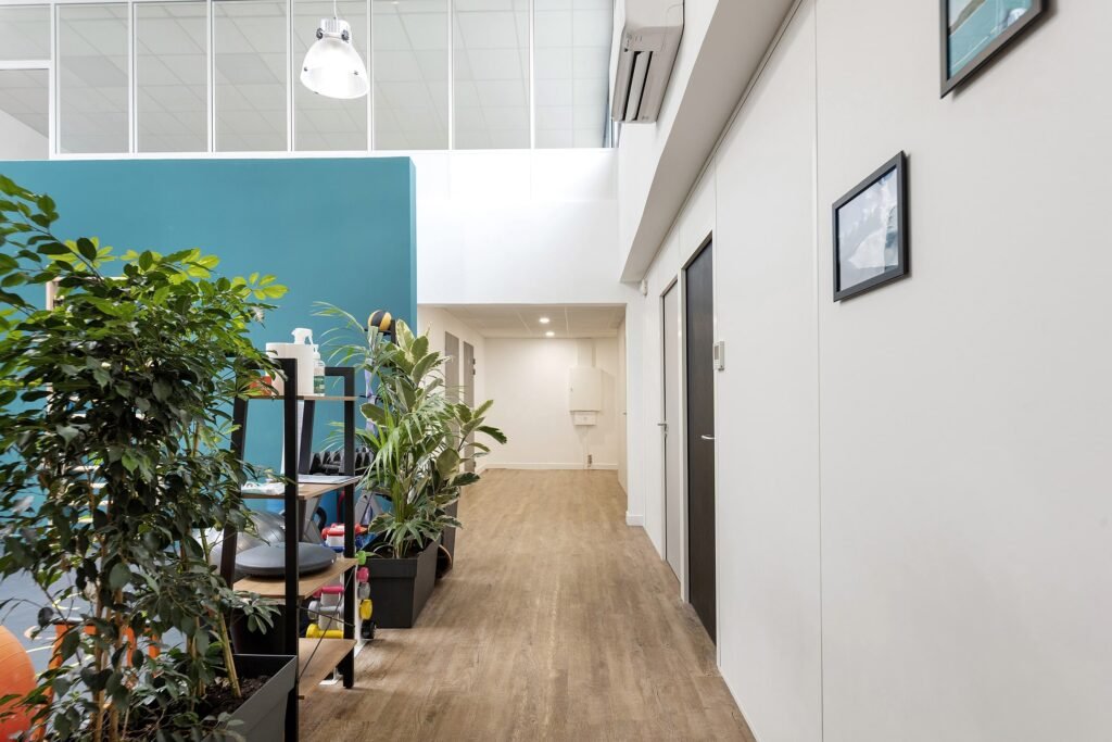 Couloir de bureau moderne avec sol en bois, plantes d'intérieur sur des étagères, mur d'accent sarcelle et murs blancs. Un tableau encadré est accroché à droite. Les plafonniers et la cloison en verre sont visibles au-dessus.
