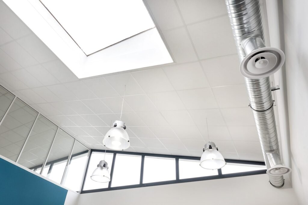 Plafond de style industriel avec un grand puits de lumière, des conduits métalliques apparents et trois dômes lumineux suspendus au-dessus d'un espace intérieur moderne aux murs blancs.