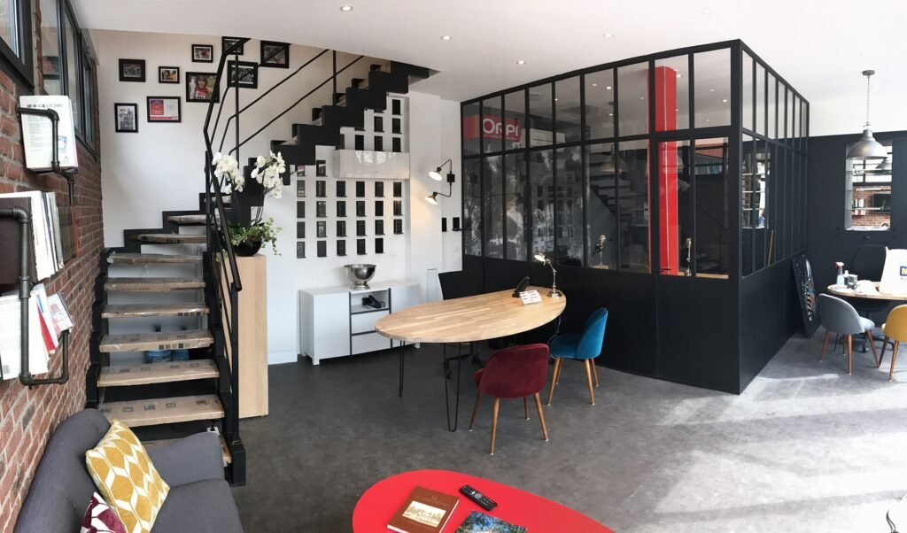 Espace de bureau moderne avec une salle de réunion vitrée, des escaliers avec des photos murales, une table ronde en bois avec des chaises colorées et divers objets de décoration.